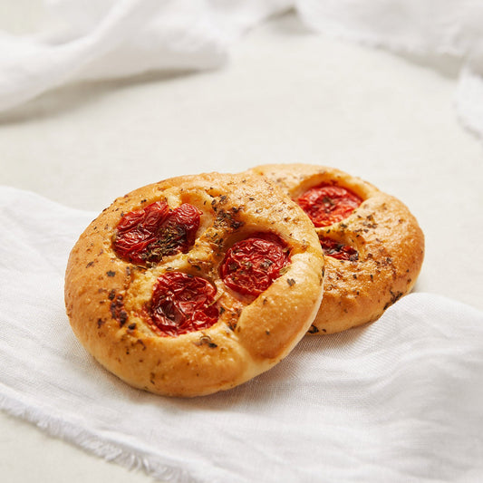 Two round focaccia bread rolls topped with sliced red tomatoes and herbs, resting on a white textured cloth against a light neutral background.