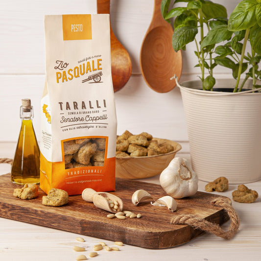 Packaging of 'Taralli al Pesto' with visible basil pesto ingredients like garlic and basil, alongside a bottle of olive oil and a bowl of Taralli crackers on a wooden board.