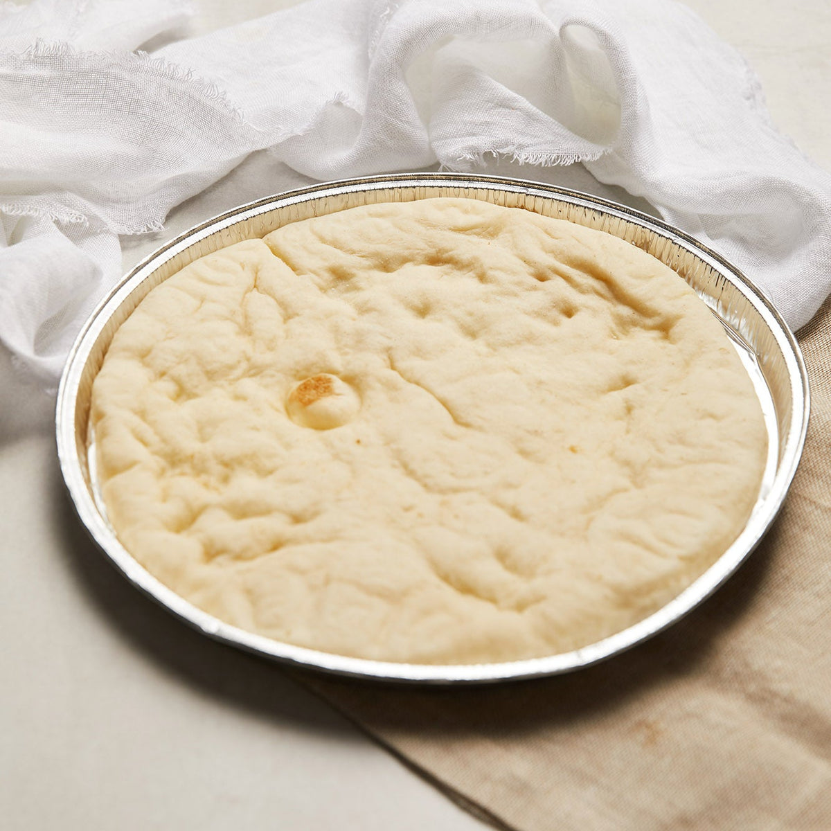A round aluminium tin containing pizza base, styled with a white cloth on a neutral beige surface.