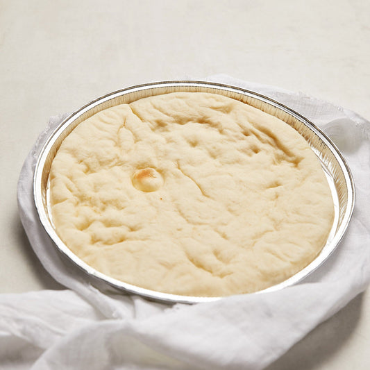 A round aluminium tin filled with a pizza base, resting on a white cloth over a light neutral background.