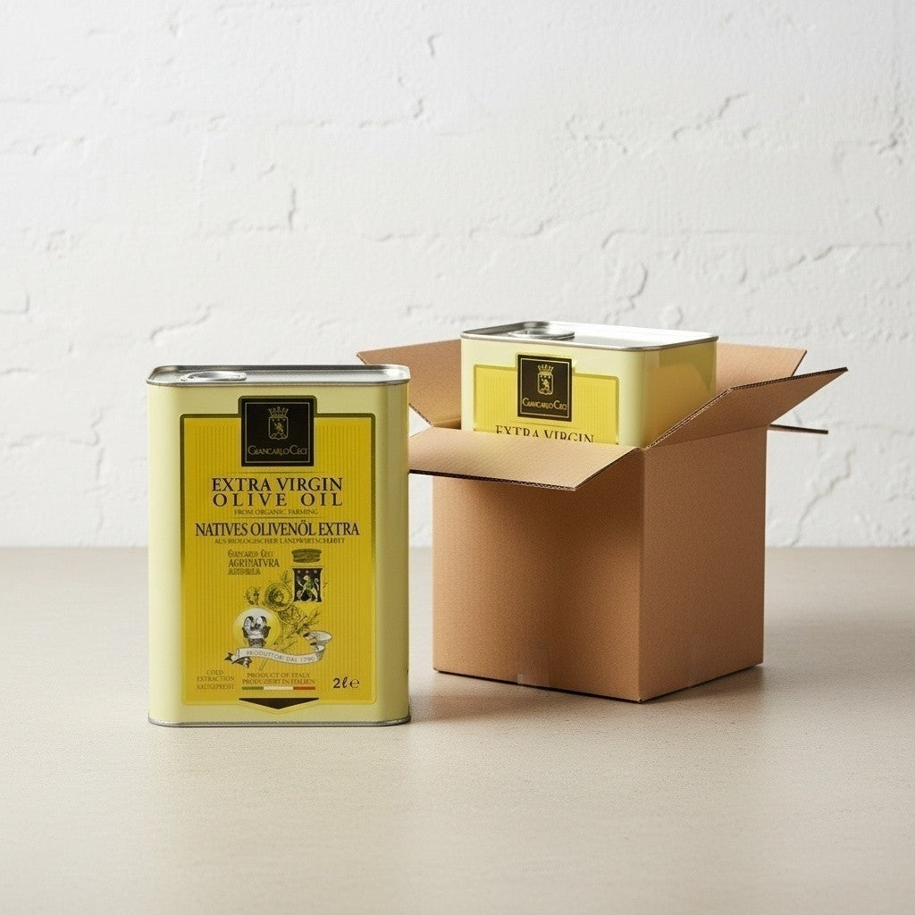 Two yellow metal tins of olive oil stand on a tabletop against a white brick wall, with one tin beside an open brown cardboard box. The labels read 'Extra Virgin Olive Oil'