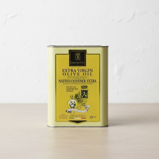 A 2-liter metal tin of Extra Virgin Olive Oil, featuring a yellow label with black text, standing on a light wooden surface against a white textured wall.