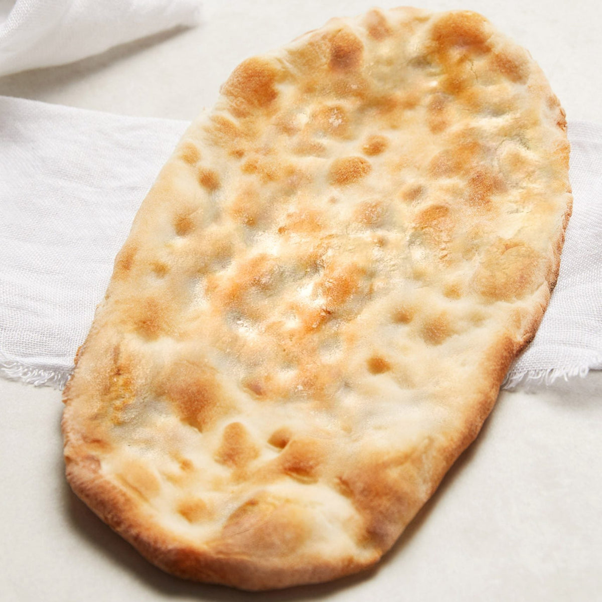 A single flatbread with a puffy, golden-brown surface, photographed on a light neutral surface with a white cloth in the background.