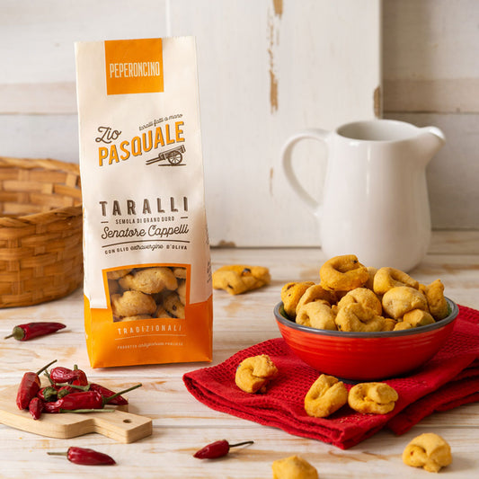 A packet of Taralli snacks on a wooden table with fresh chilli peppers and a red bowl full of taralli crackers.