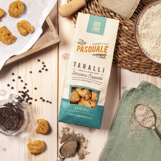 A packet of taralli on a wooden table with whole and ground pepper, placemat and kitchen towel  