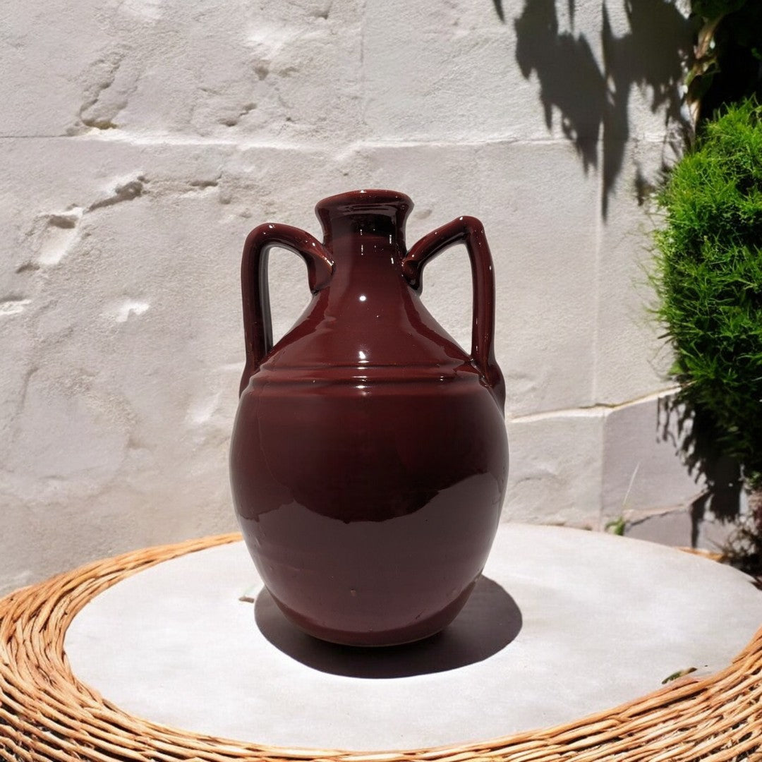 ceramic vase amphora in burgundy on a wicker table with a white stone wall and plants in the background