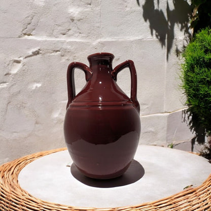 ceramic vase amphora in burgundy on a wicker table with a white stone wall and plants in the background