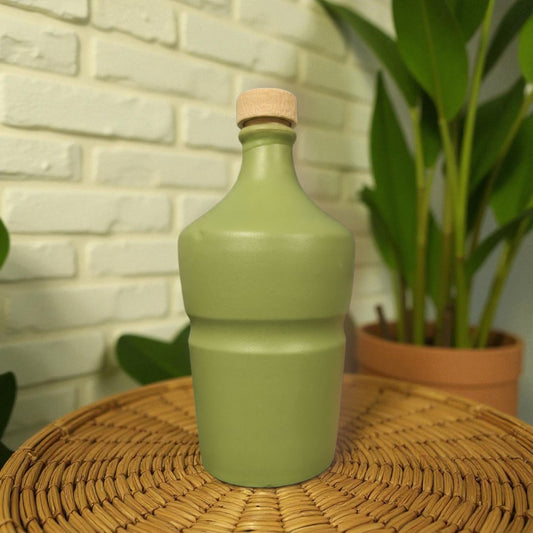 Green ceramic bottle with cork lid on a wicker table with a brick wall and plant in the background
