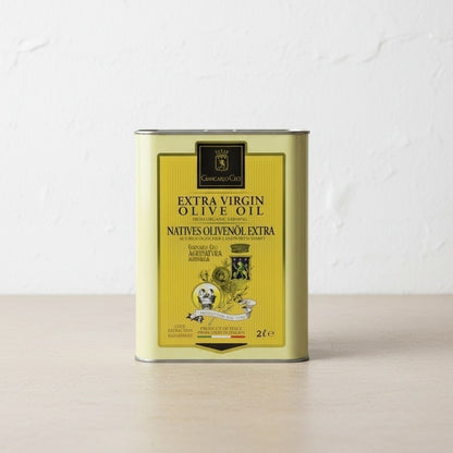 A 2-liter metal tin of Extra Virgin Olive Oil, featuring a yellow label with black text, standing on a light concrete surface against a white textured wall.