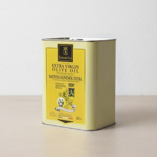 A 2L metal tin of extra virgin olive oil, featuring a yellow label with black text and illustrations, set on a light beige surface against a white textured wall.