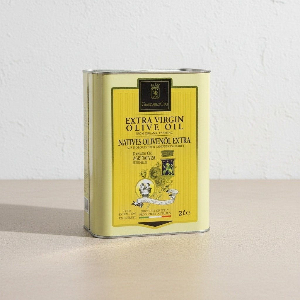 A 2-liter metal tin of Extra Virgin Olive Oil, featuring a pale yellow label with black text and illustrations, standing on a light beige surface against a white textured wall.