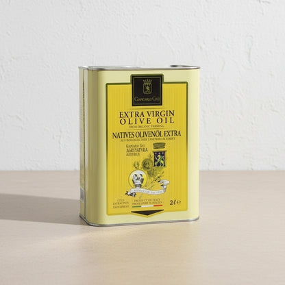 A 2-liter metal tin of Extra Virgin Olive Oil, featuring a pale yellow label with black text and illustrations, standing on a light beige surface against a white textured wall.