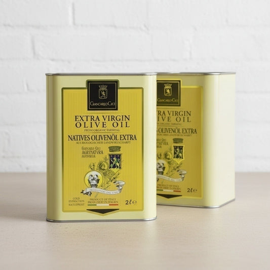Two metal tins of Extra Virgin Olive Oil on a light beige surface against a white brick wall background.
