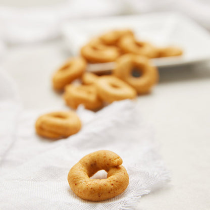 Small golden-brown ring-shaped taralli crackers arranged on a white textured cloth, with a shallow depth of field showing more taralli in the background on a white plate.