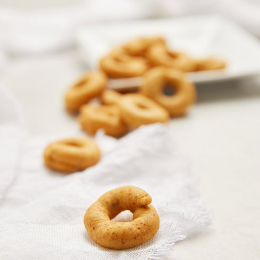 Small golden-brown ring-shaped taralli crackers arranged on a white textured cloth, with a shallow depth of field showing more taralli in the background on a white plate.